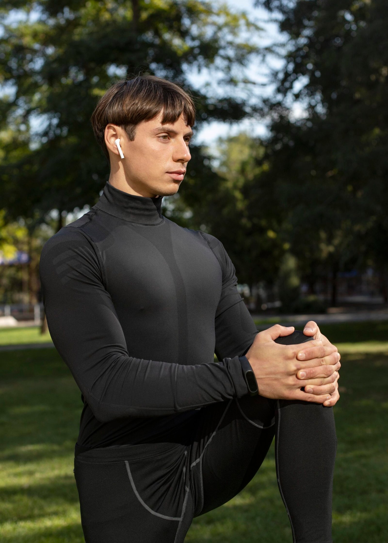 Hombre con camiseta deporte térmica de alto rendimiento y leggins largos negros estirando su pierna con un fondo de arboles y césped.