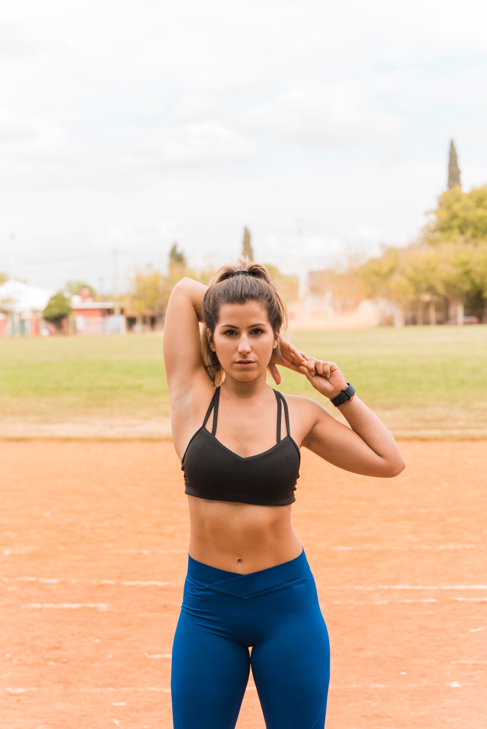 Mujer con top negro y leggins azules haciendo estiramientos en una pista de atletismo.