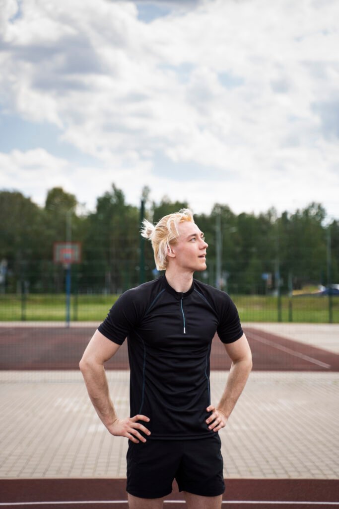 Hombre con camiseta de deporte negra y pantalones cortos de deportivos negros, con las manos apoyadas a la cintura y en una pista de pádel con arboles de fondo.