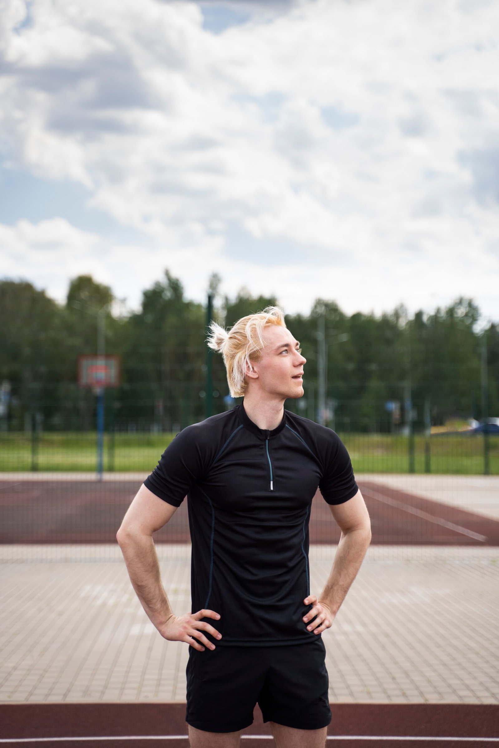 Hombre con camiseta de deporte negra y pantalones cortos de deportivos negros, con las manos apoyadas a la cintura y en una pista de pádel con arboles de fondo.